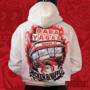 Pode incluir: Um moletom cinza claro com um design gráfico nas costas. O design apresenta as palavras "BABA YAGAS HOUSE OF CHICKEN & WAFFLES" em uma ilustração vermelha e preta. O moletom tem capuz e bolso canguru.