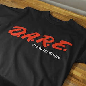 Puede incluir: Camiseta negra con la palabra "DARE" en letras rojas y la frase "me to do drugs" en letras blancas.
