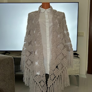 Puede incluir: Un chal beige tejido a ganchillo con un patrón floral y flecos. El chal está drapeado sobre una camisa blanca con cuello en un maniquí. El diseño de tejido abierto del chal es visible, y los flecos cuelgan.