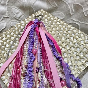 May include: A pink and purple tassel keychain with beads and ribbons. The tassel is made of various fabrics and textures, including satin, lace, and frayed fabric.