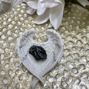 May include: A black crystal sits inside a white ceramic angel wing dish. The dish is on a gold and white patterned surface.