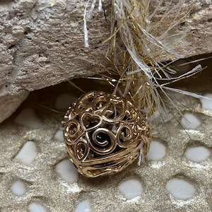 May include: A gold-colored, filigree orb pendant with a swirling design. The pendant is suspended by white and gold strands. The background is a textured, gold-colored surface.