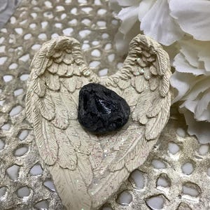 May include: A cream-colored angel wing-shaped dish with glitter accents holds a dark, textured stone. The wings are detailed with feather patterns. The background features a textured, metallic surface and white floral accents.