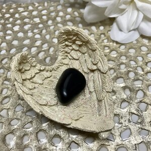 May include: A black obsidian stone resting in a white ceramic angel wing dish. The dish is decorated with glitter and has a heart shape.