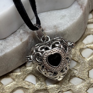 May include: A silver heart-shaped pendant with an openwork design and a black center. The pendant hangs from a black cord. The heart is embellished with small, clear stones around the inner heart shape.