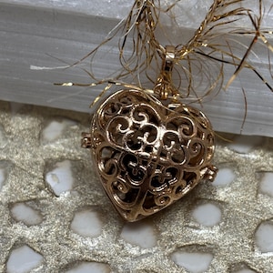 May include: A gold-colored heart-shaped pendant with an intricate filigree design. The pendant hangs from a delicate gold chain and is displayed against a textured, gold-toned surface. The heart's openwork reveals a darker interior.