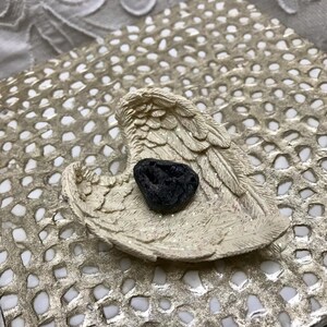 May include: A cream-colored angel wing-shaped dish with a black stone resting inside. The dish is on a gold-colored, patterned surface. The wings have detailed feathering and a glittery finish.