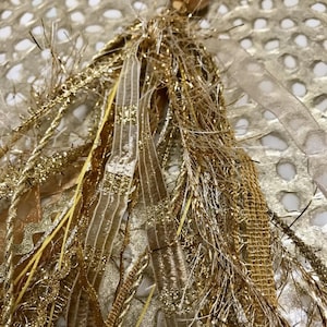 May include: A gold tassel with a variety of textures, including metallic threads, ribbons, and fringe. The tassel is hanging from a gold bead.