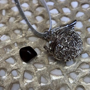 May include: A silver-colored necklace with a heart-shaped pendant and a small, dark brown stone. The pendant is a sphere with heart cutouts and small wings. The necklace is on a silver ribbon.
