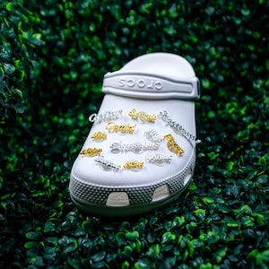 May include: White Crocs shoe with gold and silver charms featuring zodiac signs. The charms include Sagittarius, Gemini, Aquarius, Pisces, Taurus, Virgo, Libra, Cancer, Capricorn, and Scorpio.