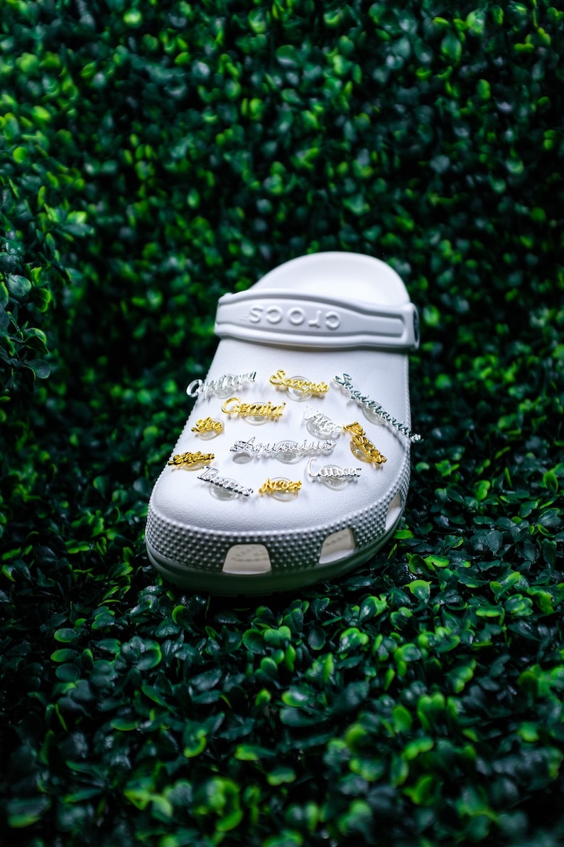 May include: White Crocs shoe with gold and silver charms featuring zodiac signs. The charms include Sagittarius, Gemini, Aquarius, Pisces, Taurus, Virgo, Libra, Cancer, Capricorn, and Scorpio.