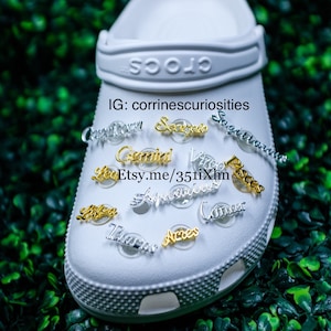 May include: White Crocs shoe with gold and silver zodiac charms attached. The charms include Capricorn, Sagittarius, Scorpio, Gemini, Virgo, Pisces, Leo, Aquarius, Cancer, Libra, Taurus, and Aries. The text on the shoe reads "Crocs" and "IG: corrinnescuriosities Etsy.me/351i1Xhn".