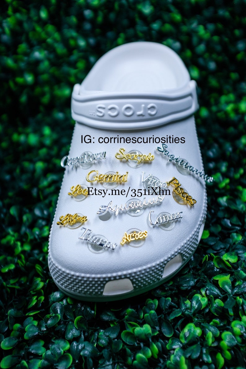 May include: White Crocs shoe with gold and silver zodiac charms attached. The charms include Capricorn, Sagittarius, Scorpio, Gemini, Virgo, Pisces, Leo, Aquarius, Cancer, Libra, Taurus, and Aries. The text on the shoe reads "Crocs" and "IG: corrinnescuriosities Etsy.me/351i1Xhn".