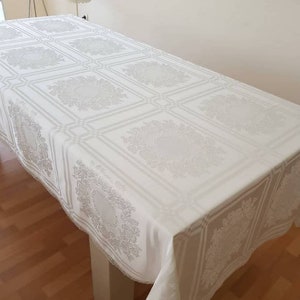 May include: White rectangular tablecloth with a delicate floral pattern. The tablecloth has a lace trim and is laid on a wooden table.