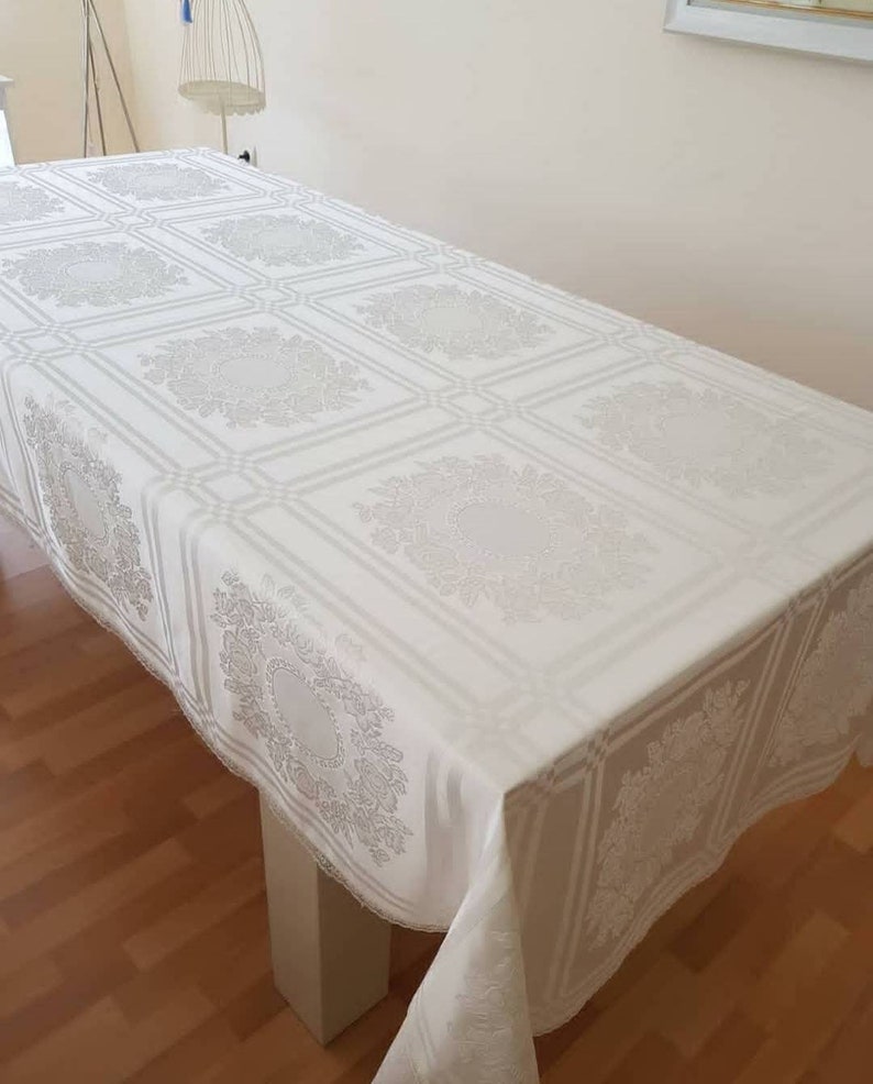 May include: White rectangular tablecloth with a delicate floral pattern. The tablecloth has a lace trim and is laid on a wooden table.
