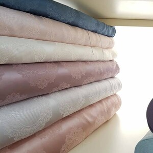 May include: A stack of fabric rolls with a delicate, intricate pattern. The rolls are in various shades of pink, white, and gray. The fabric has a subtle sheen and a soft, luxurious feel.