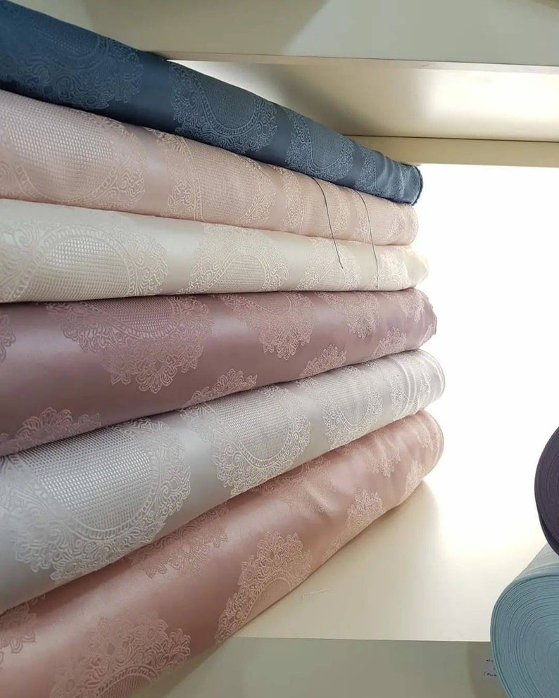 May include: A stack of fabric rolls with a delicate, intricate pattern. The rolls are in various shades of pink, white, and gray. The fabric has a subtle sheen and a soft, luxurious feel.