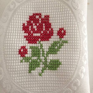 May include: White pillow with a red rose embroidered in the center. The rose is surrounded by a green stem and leaves. The pillow has a decorative border.