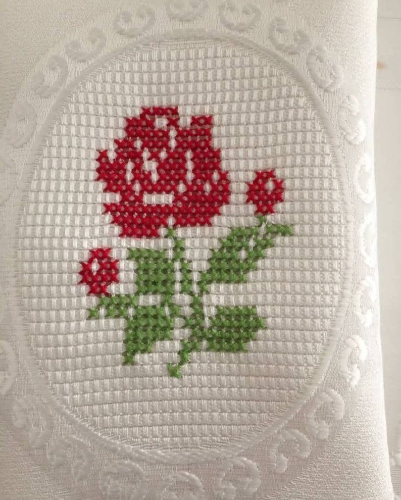 May include: White pillow with a red rose embroidered in the center. The rose is surrounded by a green stem and leaves. The pillow has a decorative border.