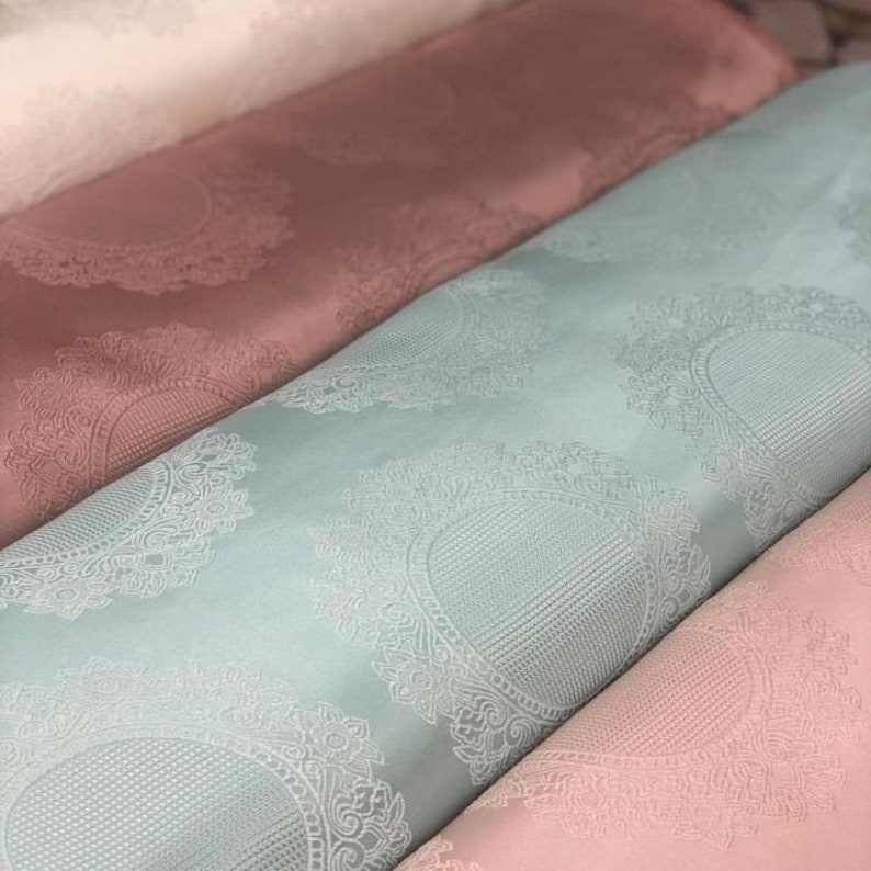 May include: Four fabric swatches in different colors, each with an embossed pattern of a circular design. The swatches are stacked on top of each other, with the white swatch on top, followed by the pink, light blue, and then the darker pink swatch.