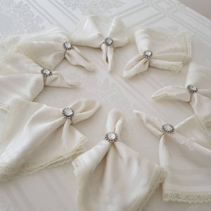 May include: White linen napkins with lace trim and jeweled napkin rings. The rings have a clear crystal stone.
