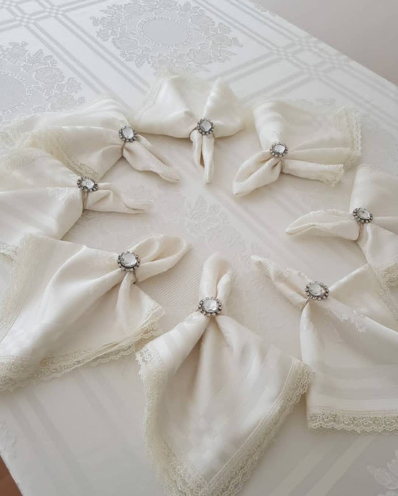May include: White linen napkins with lace trim and jeweled napkin rings. The rings have a clear crystal stone.