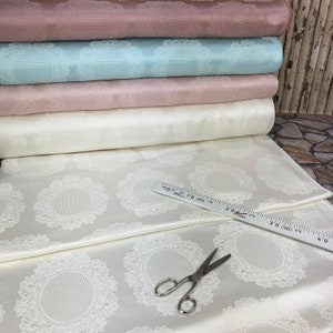 May include: A stack of white, pink, and light blue fabric with a delicate lace pattern. The fabric is folded and laid out on a white surface with a ruler and scissors.