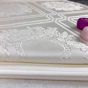 May include: A white fabric with a floral pattern and a textured surface. The fabric is folded in half and the edges are visible.