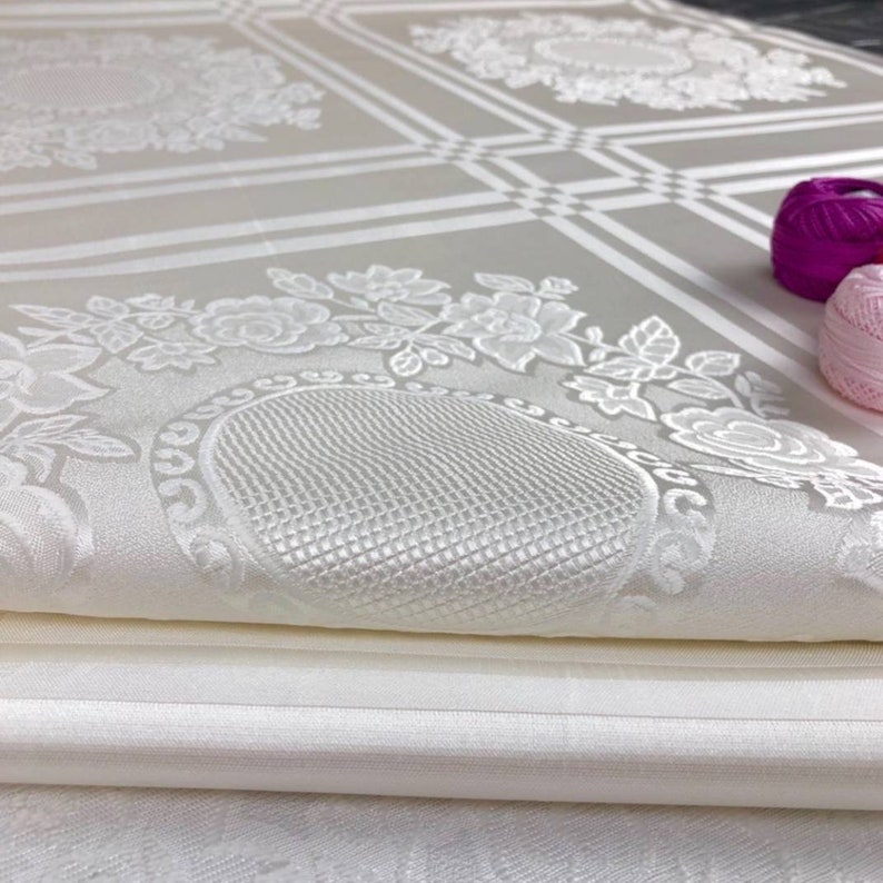 May include: A white fabric with a floral pattern and a textured surface. The fabric is folded in half and the edges are visible.