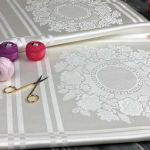May include: White fabric with a raised floral design. The fabric is folded in half, revealing a second floral design. There are three spools of thread in the image, one pink, one purple, and one red.