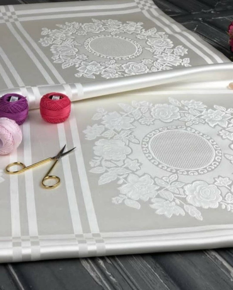 May include: White fabric with a raised floral design. The fabric is folded in half, revealing a second floral design. There are three spools of thread in the image, one pink, one purple, and one red.