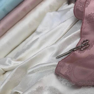 May include: A close-up of white fabric with a subtle pattern, layered over pink fabric with a more intricate pattern. A pair of silver scissors is placed on top of the fabric.