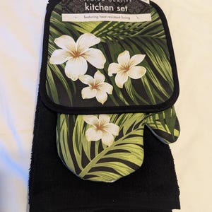 May include: A black kitchen towel with a green and white floral print pot holder and oven mitt. The pot holder and oven mitt feature a tropical leaf and white flower design. The packaging says "Premium Quality Kitchen Set featuring heat resistant lining".