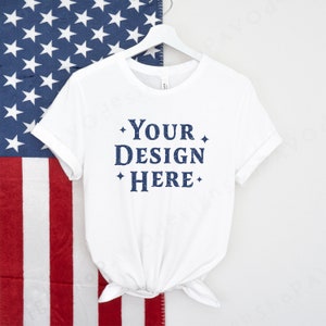 May include: White t-shirt with the text "YOUR DESIGN HERE" in blue lettering on a hanger in front of an American flag.