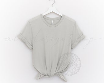 May include: A light gray t-shirt with rolled up sleeves hanging on a white hanger against a white background. The t-shirt is knotted at the bottom. A round, gray stamp with the text "Authentic Design" is visible in the lower right corner of the image.