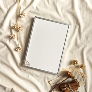 May include: A silver picture frame with a white mount on a beige fabric background. The frame is empty and ready for a photo or artwork. Dried flowers and a book are also in the image.