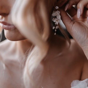Könnte beinhalten: Nahaufnahme eines Frauenohrs mit einem zarten Blumenohrring. Der Ohrring hat kleine weiße Blüten und goldene Akzente.