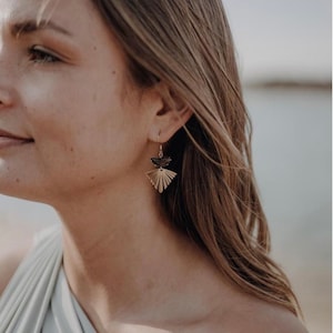 Puede incluir: Una mujer con un top azul claro de un solo hombro y pendientes dorados y negros. Los pendientes tienen un dise&ntilde;o en forma de abanico con una forma de media luna negra en la parte superior.