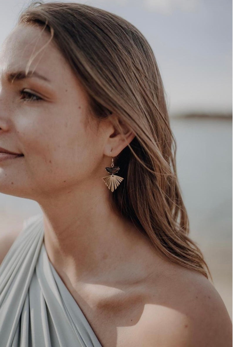 Puede incluir: Una mujer con un top azul claro de un solo hombro y pendientes dorados y negros. Los pendientes tienen un dise&ntilde;o en forma de abanico con una forma de media luna negra en la parte superior.
