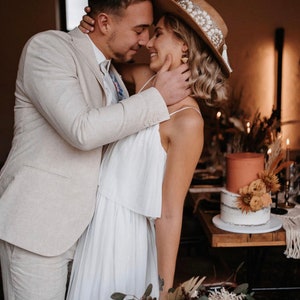 Puede incluir: Una pareja en un entorno r&uacute;stico, la novia lleva un vestido blanco y un sombrero de ala ancha con adornos, el novio lleva un traje beige claro. Se miran con afecto, la mano de la novia descansa en la mejilla del novio.
