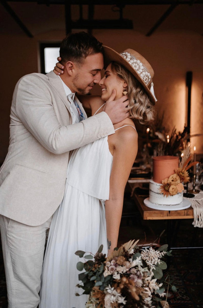 Puede incluir: Una pareja en un entorno r&uacute;stico, la novia lleva un vestido blanco y un sombrero de ala ancha con adornos, el novio lleva un traje beige claro. Se miran con afecto, la mano de la novia descansa en la mejilla del novio.