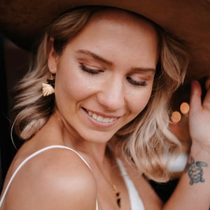 Puede incluir: Una mujer con cabello rubio que lleva un sombrero marr&oacute;n de ala ancha, un top blanco sin mangas y un collar de oro con un peque&ntilde;o colgante. Tiene un peque&ntilde;o tatuaje de tortuga en la mu&ntilde;eca.