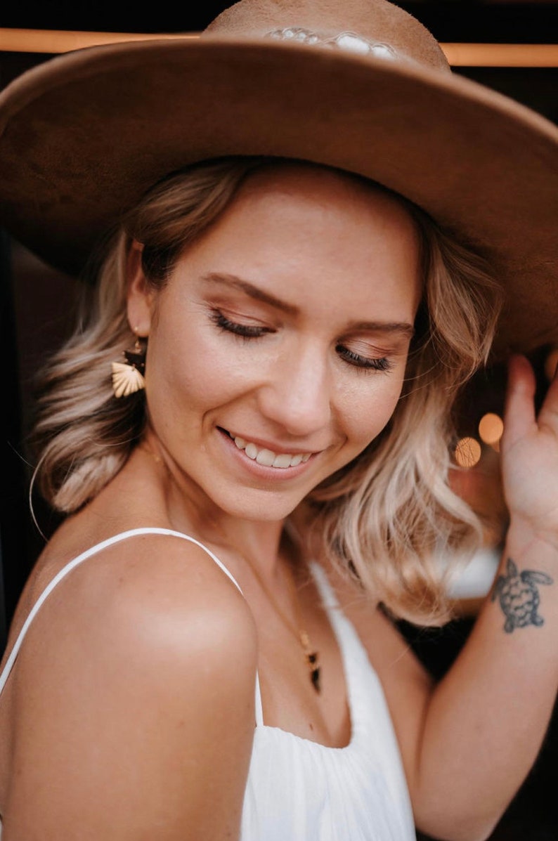 Puede incluir: Una mujer con cabello rubio que lleva un sombrero marr&oacute;n de ala ancha, un top blanco sin mangas y un collar de oro con un peque&ntilde;o colgante. Tiene un peque&ntilde;o tatuaje de tortuga en la mu&ntilde;eca.