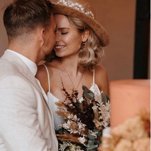 Puede incluir: Una pareja abraz&aacute;ndose, la mujer lleva un sombrero marr&oacute;n con una banda blanca y un vestido blanco, sosteniendo un ramo de flores secas. El hombre lleva una chaqueta de traje blanca.