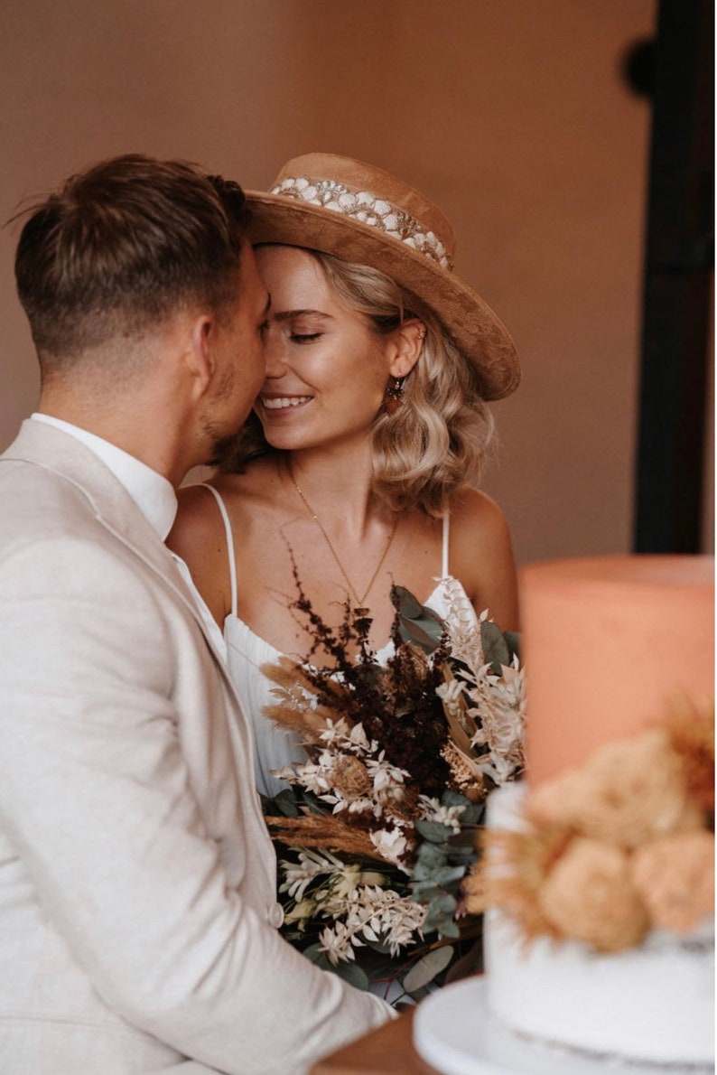 Puede incluir: Una pareja abraz&aacute;ndose, la mujer lleva un sombrero marr&oacute;n con una banda blanca y un vestido blanco, sosteniendo un ramo de flores secas. El hombre lleva una chaqueta de traje blanca.