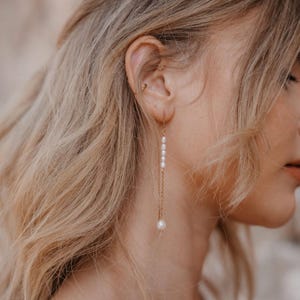 Op de afbeelding: Een lange, delicate gouden kettingoorring met kleine witte parels die eraan hangen. De oorring wordt in de onderste oorlel gedragen.