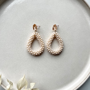Könnte beinhalten: Ein Paar cremefarbene tropfenförmige Ohrringe mit goldenen Akzenten. Die Ohrringe bestehen aus kleinen Perlen und haben einen zarten, femininen Look.