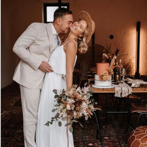 Puede incluir: Una pareja en un entorno r&uacute;stico, la novia lleva un vestido blanco y un sombrero marr&oacute;n, el novio lleva un traje beige claro, la novia lleva un ramo de flores secas.