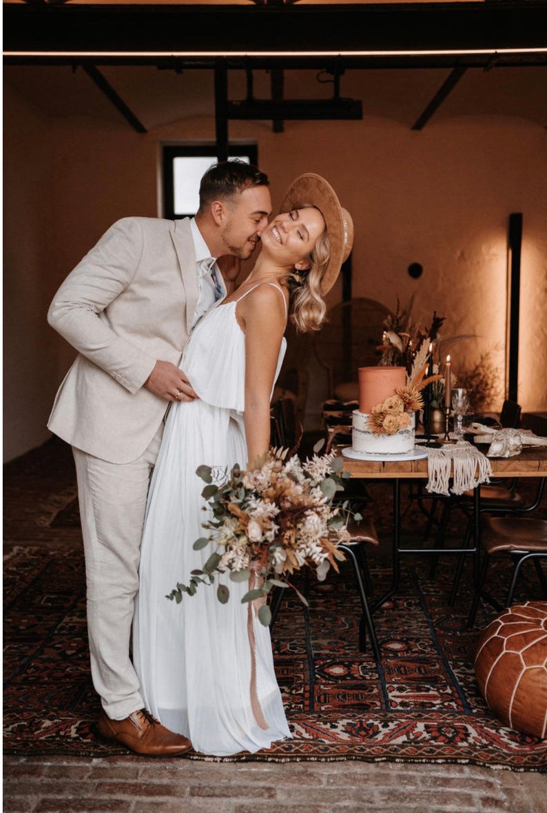 Puede incluir: Una pareja en un entorno r&uacute;stico, la novia lleva un vestido blanco y un sombrero marr&oacute;n, el novio lleva un traje beige claro, la novia lleva un ramo de flores secas.