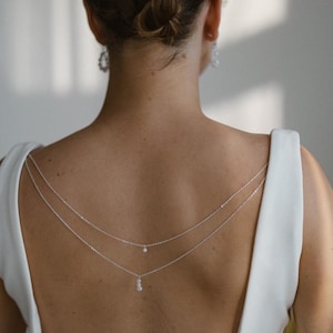 Op de afbeelding: Een zilveren rugketting met drie delicate strengen, elk versierd met kleine, sprankelende accenten. De ketting is gedrapeerd over een witte jurk, wat een elegante en minimalistische look creëert. De sieraden zijn ontworpen om op de rug te worden gedragen.